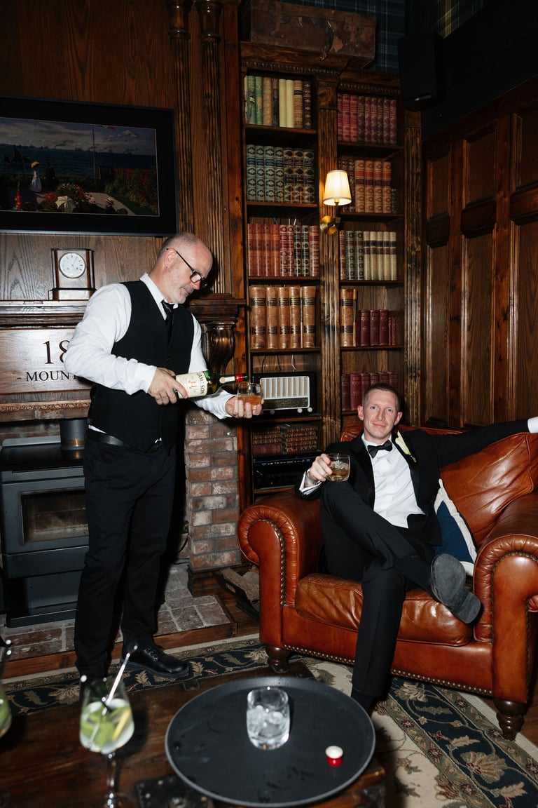 Two men in formal attire in a classic library setting. One stands pouring drinks while the other sits in a leather chair. Bookshelves and fireplace visible behind them.