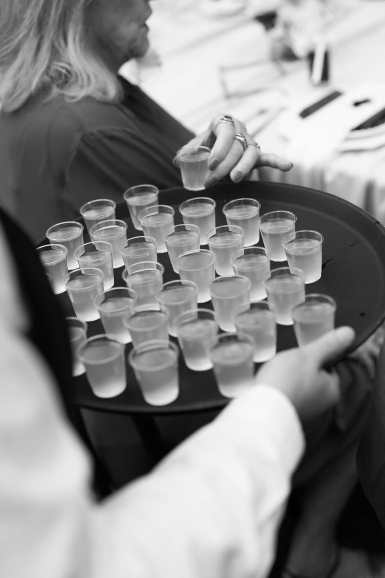 Woman holding black tray with multiple small clear glasses of water at an event, black and white photo
