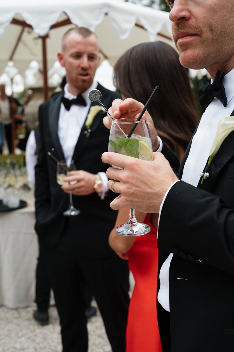 Man in tuxedo holding mojito cocktail at outdoor wedding reception with guests in formal attire in background