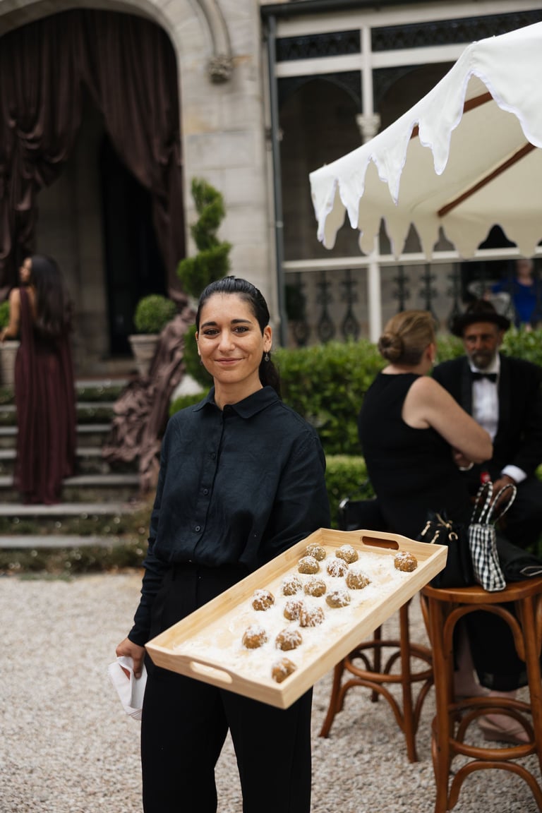 Woman in dark clothing holding a wooden tray of appetizers at an outdoor garden party with guests and white awning