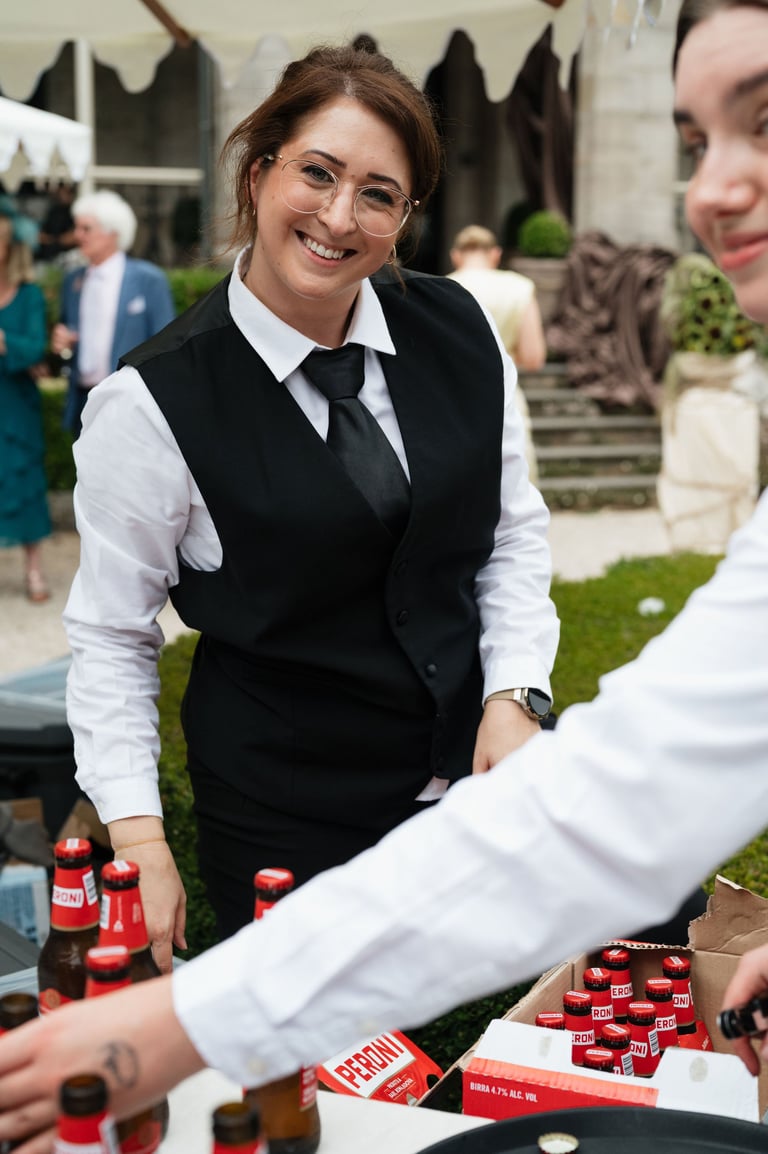 Woman in formal black vest and white shirt smiling at an outdoor event with guests and red beverages on a table
