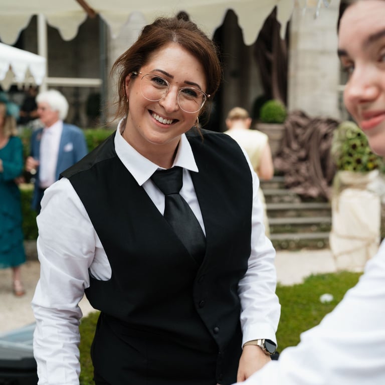 Woman in formal black vest and white shirt with glasses smiling at outdoor event with guests in background