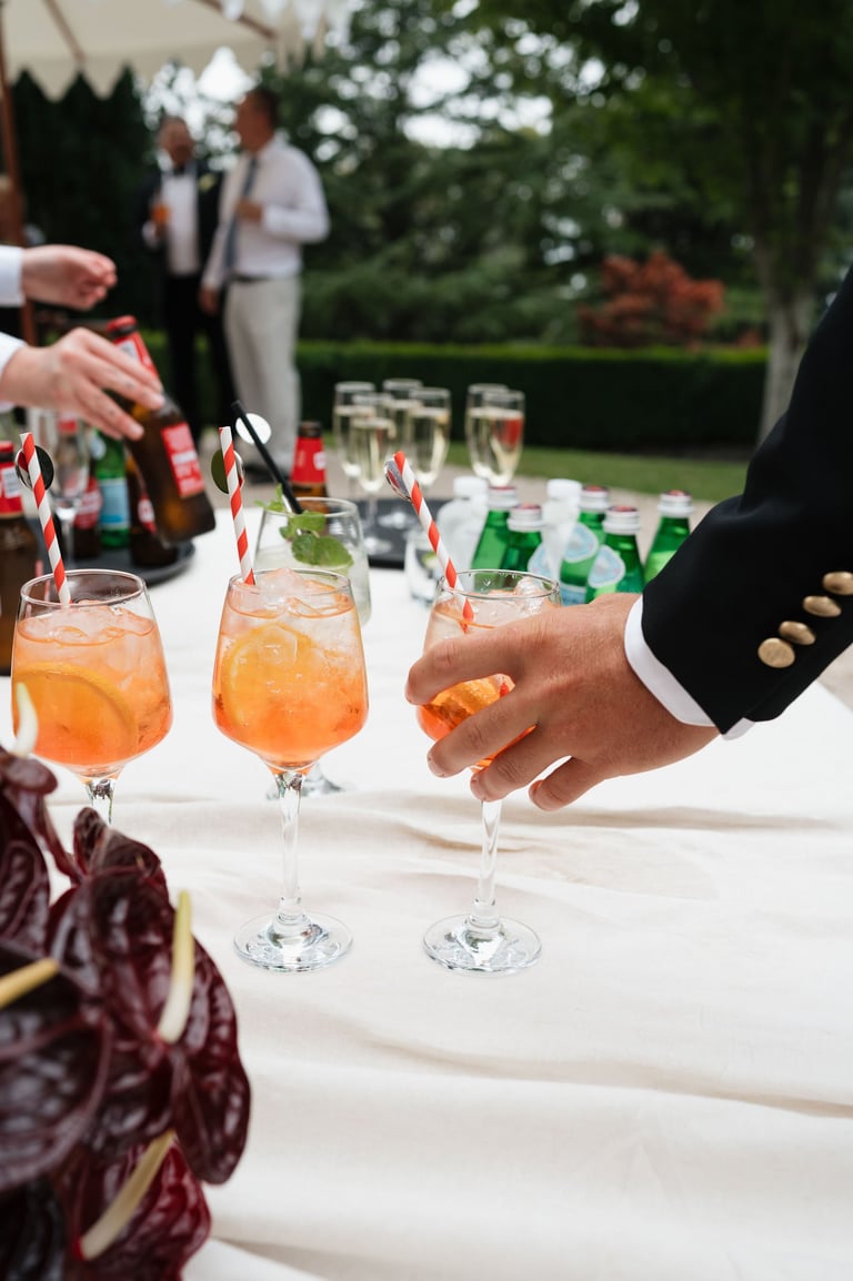 Outdoor garden party with guests holding cocktails at a white-tablecloth reception, champagne flutes with orange drinks and decorative flags