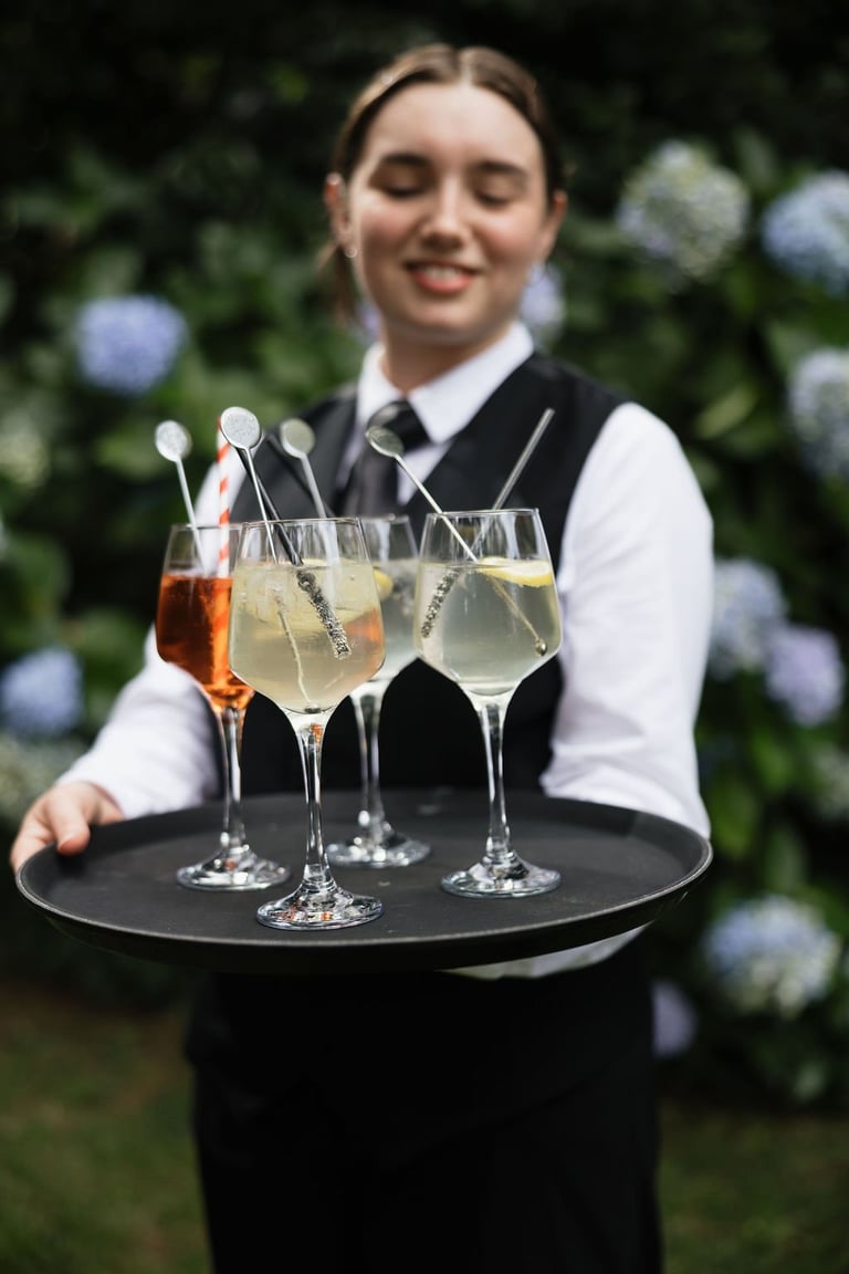 Bartender in formal attire holding a tray with three cocktails in a garden setting with blue flowers
