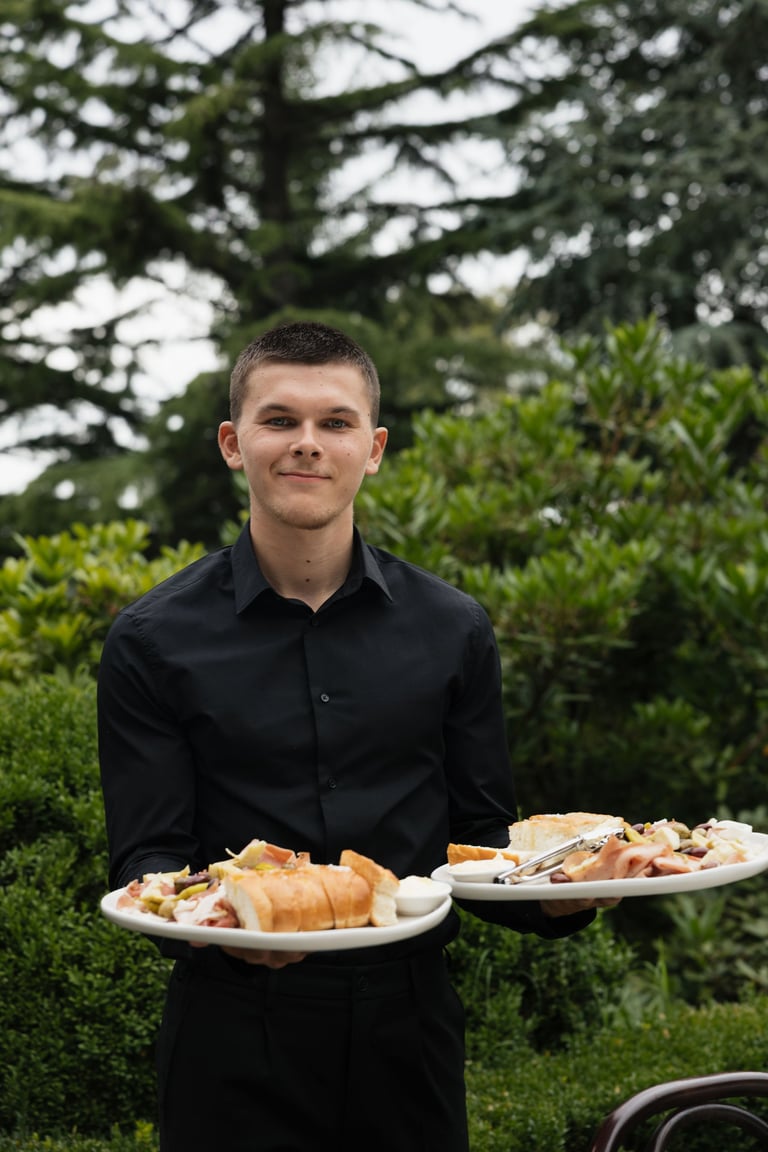 Waiter in black shirt holding two plates of food in an outdoor garden setting